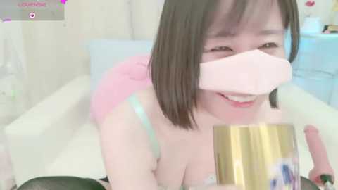 Media: A young Asian woman with straight black hair and a pale complexion, wearing a pink bra, has a pink bandage over her mouth, smiling while holding a gold-colored can. The background is a soft, pastel-colored room.