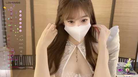 Media: Video of an East Asian woman with long brown hair, wearing a white face mask and a lace-trimmed white top, adjusting her mask in front of a wooden screen.