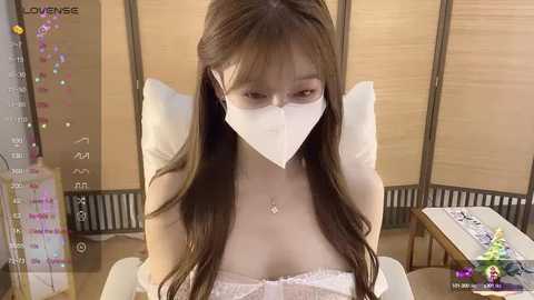 Media: Video of an Asian woman with long brown hair, wearing a white face mask, a white lace bra, and a delicate necklace, sitting in a room with wooden panels.