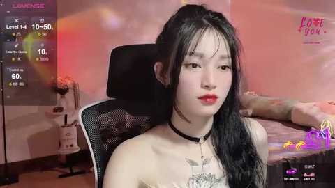 Media: Video of an Asian woman with long black hair, wearing a black choker, sitting on a black chair in a dimly lit room with a bed in the background.