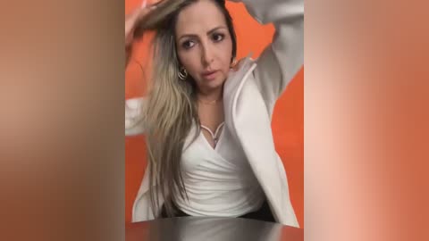 Media: Video of a woman with long, wavy, blonde hair, wearing a white blouse, sitting in a red-orange background, with blurred, out-of-focus people in the foreground.