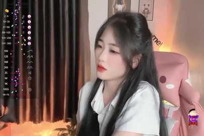 Media: A video of a young East Asian woman with long black hair, wearing a white shirt and black dress, sitting on a pink gaming chair in a cozy, dimly lit room.