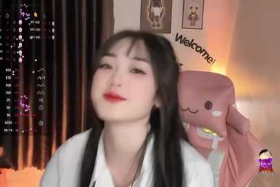 Media: A video of an Asian woman with long black hair, red lipstick, and white shirt, smiling in a dimly lit room with a pink plush toy and a \"Welcome\" sign on the wall.