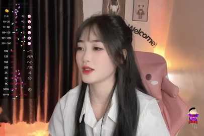 Media: Video of an East Asian woman with long black hair and pale skin, wearing a white blouse, sitting in a pink gaming chair. Background features a dimly lit room with curtains, a warm glow, and a \"Welcome to my channel\" sign.