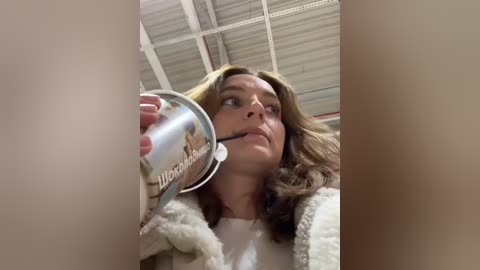 Media: Video of a young woman with long brown hair, wearing a beige furry jacket, drinking from a large, clear plastic cup, in a dimly lit room with a corrugated metal ceiling.