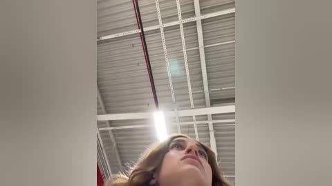 Media: A video of a young woman with wavy brown hair, looking up from a low angle, under a metal ceiling with visible beams and a single bright light bulb.