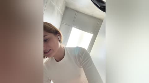 Media: Video of a light-skinned woman with short brown hair, wearing a white long-sleeve shirt, smiling as she peeks through a window.