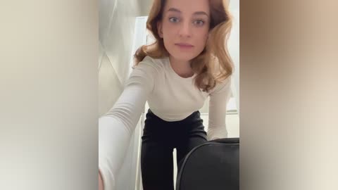 Media: A video of a young woman with wavy brown hair, wearing a white long-sleeved top and black pants, leaning forward in a narrow hallway, with beige walls and a black chair visible in the background.