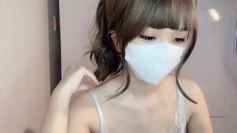 Media: Video of a young Asian woman with light skin, straight brown hair, and bangs, wearing a white surgical mask and a white lace camisole, adjusting her hair in a bathroom with beige walls.