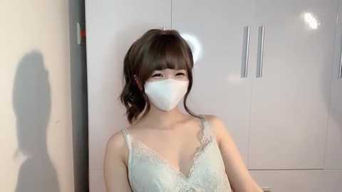 Media: Video of an East Asian woman with medium skin tone, wearing a white face mask, a white lace bra, and a ponytail. She stands against a white wardrobe with silver handles. The background includes a shadow on the left wall.