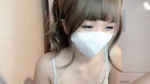 Media: Video of a young East Asian woman with fair skin, straight brown hair, wearing a white surgical mask, and a light gray lace bra. She is indoors, with a white door and beige wall in the background.