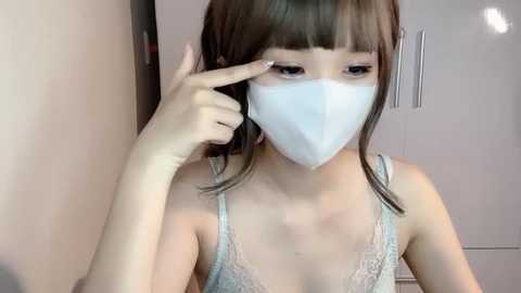 Media: Video of a young East Asian woman with straight, shoulder-length brown hair, wearing a white face mask and light blue lace lingerie. She is indoors, possibly in a bathroom, pointing at her face with her right index finger.