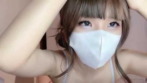 Media: Video of an East Asian woman with straight, light brown hair and bangs, wearing a white face mask, and a white tank top, adjusting her hair with both hands. Soft, pinkish background.