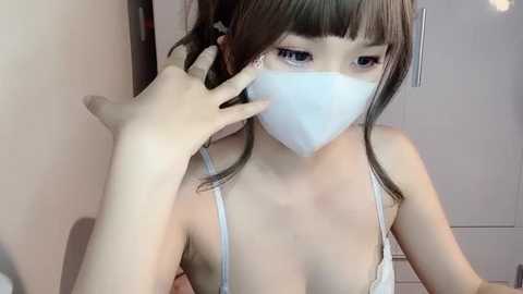 Media: Video of an Asian woman with light skin, wearing a white face mask and a white lace bra, adjusting her hair in a mirror. She has straight, dark brown hair with blunt bangs. Background shows a neutral-colored room with a white cabinet.