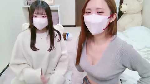 Media: Video of two young East Asian women in white masks and gray sweaters, sitting on a bed with white sheets and plush toys in the background.