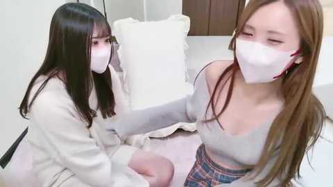 Media: Video of two young East Asian women, wearing white masks, sitting on a bed with white pillows. One has long straight black hair, the other has long straight brown hair. They wear casual clothing, including a grey top and plaid skirt.