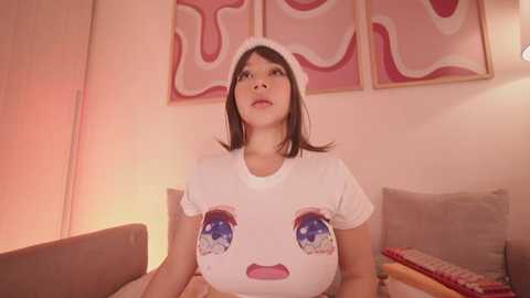 Media: A video of a young woman with shoulder-length brown hair, wearing a white t-shirt with cartoonish eye designs, sitting on a beige couch in a warmly lit room.