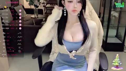 Media: Video of an Asian woman with long black hair, wearing a revealing light blue dress, seated indoors. Background includes modern furniture and a Christmas tree.