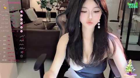 Media: Video of an East Asian woman with long black hair, wearing a black dress, seated indoors. Background shows a modern living room with a sofa and potted plant.