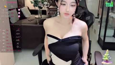 Media: Video of a young Asian woman with fair skin and braided hair, wearing a black and white off-shoulder dress, sitting in a black gaming chair in a modern living room.