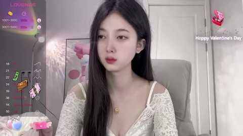 Media: Video of a young Asian woman with long black hair, wearing a white lace dress, sitting in a room with a Valentine's Day theme, including a door with a heart-shaped sticker and a pink flower.