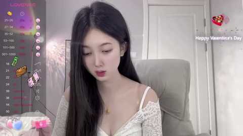 Media: A video of an East Asian woman with long black hair and fair skin, wearing a white lace off-shoulder top, sitting in a chair. The background shows a white door and a blurred pink object. The image has a digital overlay with Valentine's Day-themed icons and text.