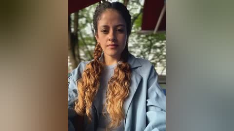 Media: Video of a young woman with light brown skin, long wavy hair, and two braided ponytails, wearing a light blue jacket, seated indoors with a blurred greenery background.