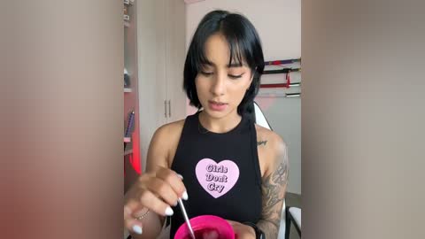 Media: Video of a young Latina woman with short black hair, tattoos, wearing a black sleeveless shirt with \"Baby's Due Date\" heart print, sitting in a modern kitchen, holding a pink bowl.