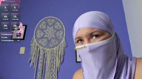 Media: A video featuring a woman in a light purple hijab and headscarf, standing in front of a purple wall adorned with a dreamcatcher and a framed photo.