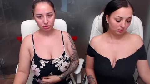 Media: Video of two women with tattoos; one in a black floral dress, the other in a black off-shoulder top. Both seated in a medical examination room with a fogged mirror and blurred background.