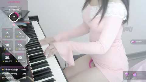 Media: A video of an Asian woman with long black hair, wearing a pink lace top and matching skirt, playing a black and white keyboard. The background is a minimalist white room.