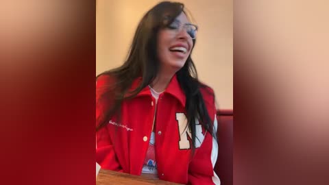 Media: Video of a joyful woman with long black hair, wearing a red blazer, smiling widely, seated indoors with blurred background.