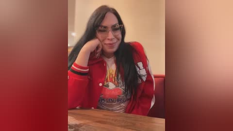 Media: A video of a person with long black hair, wearing glasses, a red jacket, and a graphic t-shirt, sitting at a wooden table in a warm-toned indoor setting.