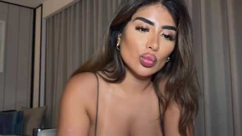 Media: Video of a curvaceous Latina woman with long, wavy brown hair, wearing a black spaghetti strap top, puckering her lips in a playful manner. She has dark, well-defined eyebrows and wears large hoop earrings.