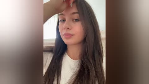 Media: A close-up video of a young woman with long, straight brown hair, fair skin, and dark eyes. She has a neutral expression, wearing a white shirt, and is standing indoors with blurred background.