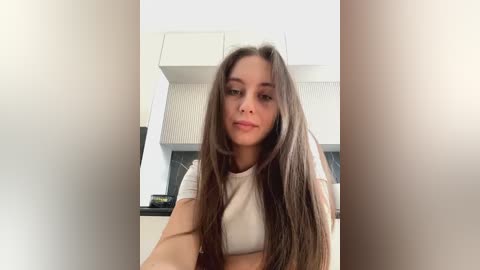 Media: A video of a young woman with long, straight brown hair, fair skin, and a slender build, wearing a white crop top, standing in a modern kitchen with white cabinets and a black countertop.