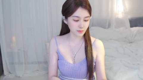 Media: Video of an East Asian woman with fair skin, long brown hair, and a slender build. She wears a light purple lace camisole and silver earrings, seated on a white bed with sheer curtains in the background.