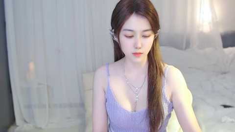 Media: A video of an East Asian woman with fair skin, long brown hair, wearing a light purple lace dress, sitting on a bed with white linens and sheer curtains in the background.