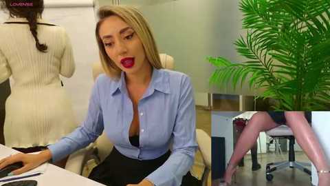 Media: Video: Blonde woman with red lipstick, blue blouse, and black skirt sits at desk. Background shows a mannequin in a white sweater and another woman in a black dress.