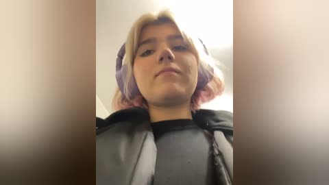 Media: Video of a young woman with shoulder-length, wavy, pink hair, wearing a black leather jacket and headphones, taken from a low angle. The background is blurred, suggesting an indoor setting.