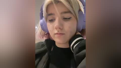 Media: A video of a young girl with fair skin and blonde hair, wearing purple headphones and a black jacket, looking introspective.