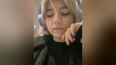 Media: Video of a young woman with fair skin, light blonde hair, wearing large purple headphones, resting her chin on her hand. She is dressed in a black top, with a blurred background.