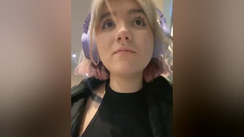 Media: Video of a young woman with light skin, wearing pastel-pink bobbed hair and large purple headphones. She has a neutral expression and is dressed in a black top. The background is blurred, suggesting an indoor setting.