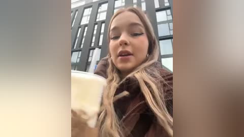 Media: A close-up video of a young woman with long, wavy blonde hair, wearing a brown plaid coat, sipping a frothy beer in an urban setting with modern glass buildings in the background.