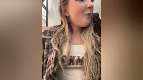 Media: Video of a young Caucasian woman with long, wavy blonde hair, wearing a brown and white plaid shirt and white tank top. She has fair skin, makeup, and a slight smile. Background shows a blurred indoor setting.