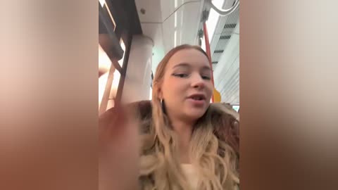 Media: A candid video captures a young woman with fair skin, long blonde hair, and a fur coat, looking out of a subway car window. The background features modern subway decor with white walls and metallic elements.