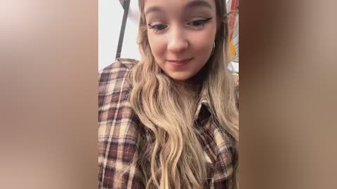 Media: Video of a young woman with long, wavy blonde hair, wearing a plaid shirt, smiling slightly. Background is blurred, featuring a white wall and indistinct objects.