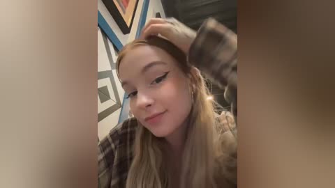 Media: Video of a young woman with fair skin and long, straight blonde hair, wearing a plaid shirt, sitting on a bed with geometric-patterned wallpaper in the background.