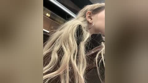 Media: Video of a blonde woman with long, wavy hair, wearing a brown jacket, seen from the side. Background shows a blurred indoor setting with metallic accents.