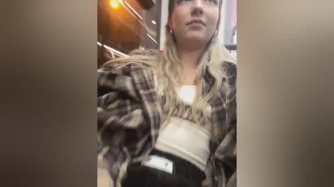 Media: Video of a young Caucasian woman with long, wavy blonde hair, wearing a plaid shirt over a white tank top and black high-waisted pants, standing in front of a blurred, lit-up store window.